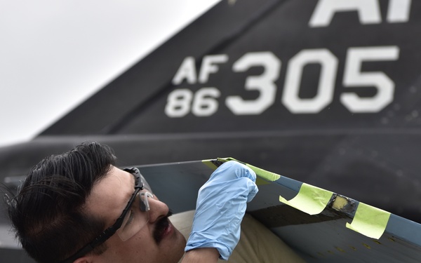 A military maintenance technician is performing repair or inspection work on a military aircraft, visible by the tail section marked 'AF 86-305' with 'AK' tail code. The technician is wearing blue nitrile gloves and safety glasses while applying or inspecting what appears to be masking tape (yellow/green) on a control surface or panel edge. The setting appears to be an outdoor flight line or maintenance area under overcast skies.