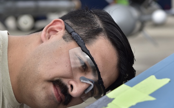 A man wearing protective safety goggles and a light-colored t-shirt is closely inspecting or working on a blue painted surface. The surface has yellow masking tape applied, suggesting a painting or surface repair operation is underway. The setting appears to be an outdoor tarmac or maintenance area based on the background.
