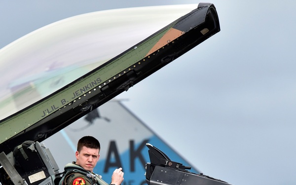 A military pilot is seated in the open cockpit of a fighter jet with a green and brown camouflage paint scheme. The canopy is raised and the pilot appears to be preparing for or completing a flight operation. The aircraft displays visible rescue markings, safety instructions, and the tail code 'AK', consistent with a U.S. Air Force F-16 Fighting Falcon.