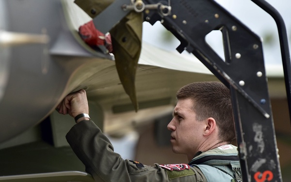 A military pilot or crew member in a flight suit and harness is conducting a pre-flight inspection of a military jet aircraft. The individual is closely examining the aircraft's intake or underside section, suggesting a routine walk-around check before flight operations. A ground support ladder labeled 'SPARE 1' is visible in the background.