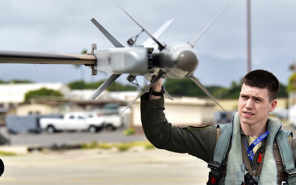 A military pilot in full flight gear conducts a pre-flight or post-flight inspection of an aircraft on a tarmac. He is touching or examining what appears to be an air-to-air missile mounted on the aircraft's wing pylon. The setting appears to be a military airfield, likely in a tropical or subtropical location based on the vegetation and architecture visible in the background.