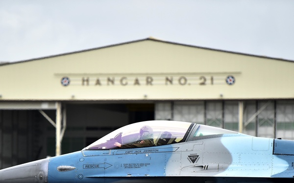 A blue camouflage-painted F-16 Fighting Falcon jet fighter taxis on the flight line in front of Hangar No. 21 at a military air base. The aircraft bears the number '22' in red on its fuselage and is named 'CAPT JACK JOHNSTON' below the cockpit. A pilot is visible in the cockpit wearing a flight helmet.