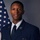 A formal military portrait of a United States Air Force enlisted member in dress blue uniform, posed in front of an American flag against a gray studio background. The subject displays rank insignia consistent with Senior Airman (E-4) on the left sleeve, along with service ribbons and a badge on the chest. This appears to be an official military service photograph.