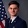 A formal military portrait of a United States Air Force enlisted member in dress blues uniform, posed in front of an American flag against a gray studio background. The subject displays several ribbons, a badge, and rank insignia consistent with the grade of Staff Sergeant. This appears to be an official service portrait.
