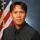 A formal portrait photograph of an adult male wearing a dark gray vest over a black dress shirt with a black tie. The subject is posed against a studio blue-gray background with a United States flag visible to the left. The image appears to be an official or professional headshot.