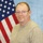 A middle-aged man poses for what appears to be an official or professional portrait photograph. He is positioned in front of an American flag and a light blue backdrop, suggesting a government, military, or civic organization setting. He wears a gray knit sweater and thin-framed rectangular glasses.