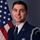 A formal military portrait of a young male U.S. Air Force Staff Sergeant in dress blues uniform. He is a member of the Base Honor Guard, as indicated by multiple insignia and a shoulder patch. The portrait is taken against a gray studio background with a U.S. flag visible to the left.