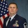 This is a formal military portrait of a U.S. Air Force Senior Master Sergeant in dress blues uniform. The subject is posed in front of an American flag against a standard blue studio backdrop, consistent with official military photography. Multiple service ribbons, badges, and rank insignia are clearly visible on the uniform.