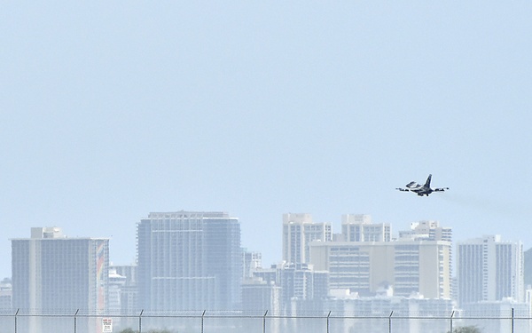 A military or commercial jet aircraft is captured in flight over a coastal airport runway, with a dense urban skyline visible in the background through atmospheric haze. The foreground features turquoise water and a rocky seawall or breakwater, suggesting the airport is situated on or near a coastal peninsula. The scene appears to be taken from across a body of water looking toward the airfield and city.
