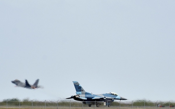 The image shows a military airfield situated adjacent to a body of water, with one F-16 Fighting Falcon jet taxiing or stationary on the runway and a second aircraft appearing to take off or bank steeply in the background. The stationary aircraft displays 'AK' tail markings, indicating it belongs to an Alaska Air National Guard unit. A rocky seawall separates the airfield from the turquoise water in the foreground.
