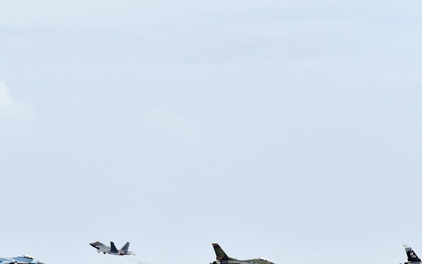 The image shows a coastal military airfield with four fighter jets visible along the runway. One aircraft appears to be in the process of taking off or landing, while the others are positioned on the tarmac. The scene is viewed from across a body of turquoise water, with a rocky seawall separating the water from the airfield.