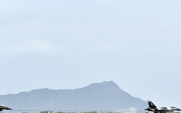 Two military fighter jets, consistent with F-16 Fighting Falcons, are taxiing or taking off on a runway situated on a narrow strip of land surrounded by turquoise water. A distinctive volcanic mountain peak, consistent with Diamond Head crater, is visible in the background, suggesting this is Joint Base Pearl Harbor-Hickam in Honolulu, Hawaii. The scene is photographed from across the water, providing a wide perspective of the coastal airfield.