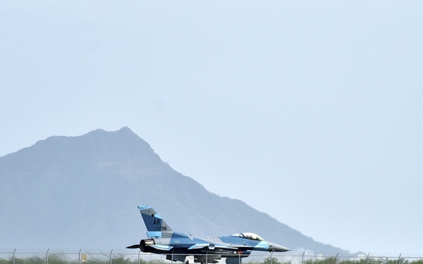 A military fighter jet, painted in a blue camouflage scheme, is taxiing on a runway at a coastal airbase. The aircraft displays 'AK' tail markings, indicating Alaska Air National Guard affiliation. The scene is set against a backdrop of turquoise water, a rocky seawall, and a prominent mountain peak under a hazy sky.