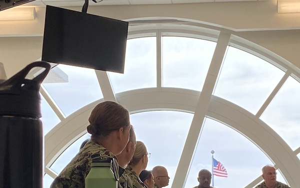 A group of U.S. Navy personnel in digital camouflage uniforms (NWU Type III) are seated at tables in what appears to be a briefing or meeting room. The room features a large arched window through which an American flag is visible outside. A CamelBak Eddy water bottle is prominently visible in the foreground.