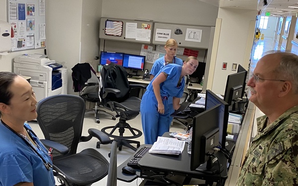 A military officer in U.S. Navy digital camouflage uniform is conversing with medical staff in blue scrubs at what appears to be a hospital nursing station. The setting is a modern clinical workspace with multiple computer workstations, monitors, and office equipment. Three healthcare workers in scrubs are visible, suggesting this may be an official visit or inspection of a military medical facility.