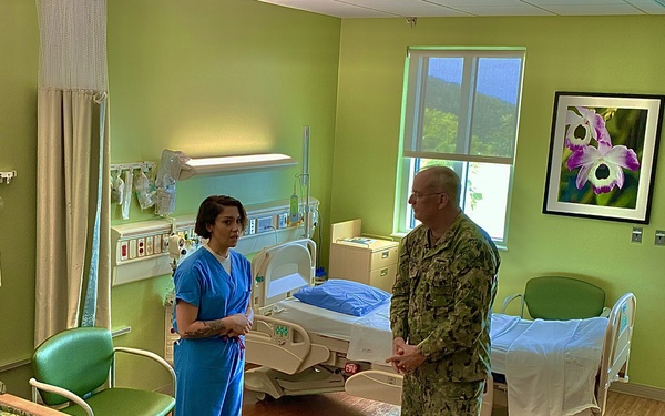 A healthcare worker in blue scrubs is conversing with a military personnel member wearing camouflage uniform inside a hospital patient room. The room appears to be unoccupied and well-maintained, featuring two hospital beds, medical equipment, and green-painted walls. The setting suggests an official visit or inspection of a military or civilian medical facility.
