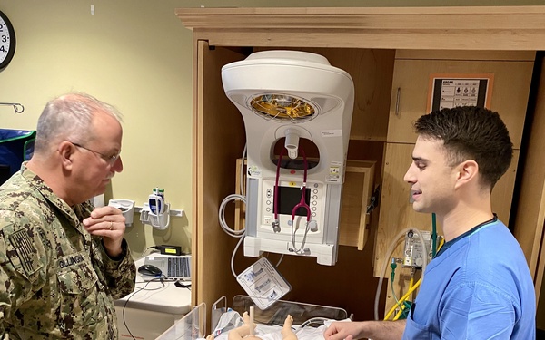 A U.S. Navy officer in camouflage uniform and a healthcare worker in blue scrubs are examining a neonatal simulation mannequin on a warmer station in what appears to be a medical simulation training room. The scene depicts a training or demonstration environment with realistic medical equipment. The Navy officer's name tape reads 'GILLINGHAM' and they appear to be receiving a briefing or demonstration from the medical professional.