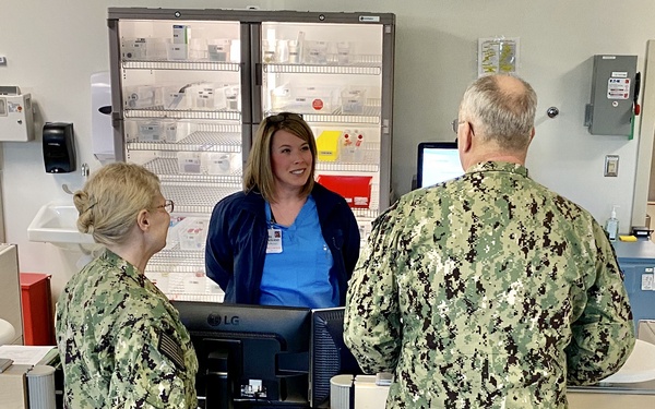 Two U.S. Navy personnel in NWU Type III (woodland digital camouflage) uniforms are standing at a medical or pharmacy counter speaking with a civilian healthcare worker in blue scrubs. The setting appears to be a clinical or hospital facility, likely a military or military-affiliated medical installation. The interaction suggests an administrative or informational exchange at a nursing station or pharmacy window.
