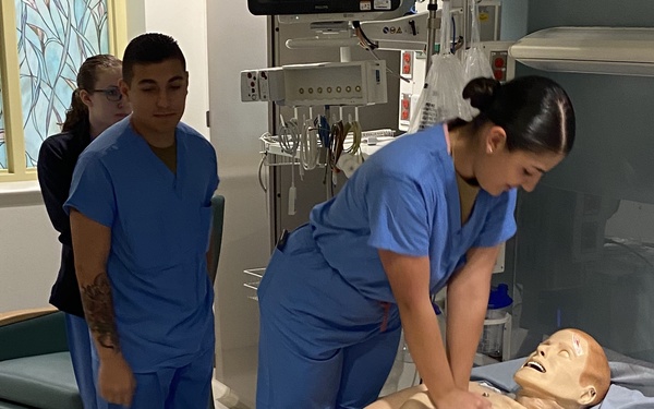 A nursing or medical training simulation is taking place in a clinical skills lab. A student in blue scrubs is performing chest compressions (CPR) on a full-body medical mannequin lying on a hospital bed. Several other students in scrubs observe the procedure, likely as part of a structured educational exercise.
