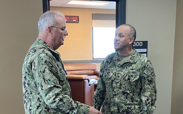 Two U.S. Navy personnel in NWU Type III (digital camouflage) uniforms are shaking hands inside what appears to be a military office building. The individual on the right has a visible name tape reading 'MENONI' and a 'U.S. NAVY' tape, consistent with the door placard behind them reading 'RADML JOHN MENONI'. They are standing on a decorative floor rug bearing what appears to be the seal of Naval Forces Marianas or a related Guam-based command.