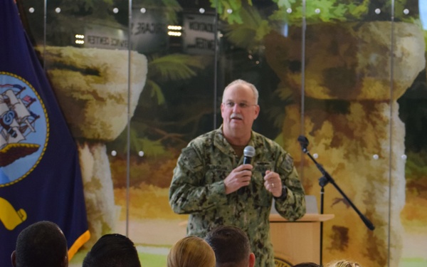 A senior military officer in U.S. Navy camouflage (NWU Type III) uniform is addressing an audience of seated military personnel at what appears to be U.S. Naval Hospital Guam. The speaker stands at a podium holding a microphone, with a U.S. Navy flag visible to the left. The audience members are wearing standard Navy Working Uniforms and at least one person is wearing a 'U.S. Naval Hospital Guam - Service Before Self' sweatshirt.