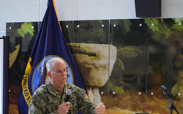 A U.S. Navy officer in digital camouflage (NWU Type III) uniform is speaking at a podium bearing the seal of U.S. Naval Hospital Guam, established 1899, with the motto 'Service Before Self.' The officer, whose name tag reads 'Gillingham,' is holding a microphone and gesturing while addressing an audience. A United States Navy flag is visible in the background.