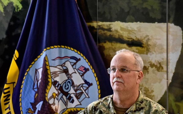 A U.S. Navy officer in NWU Type III camouflage uniform is speaking at a podium while holding a microphone. The name tape on his uniform reads 'GILLINGHAM' and the branch tape reads 'U.S. NAVY'. He is standing in front of an official United States Navy flag with the Navy seal.