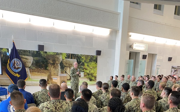 A military briefing or all-hands meeting is taking place at what appears to be U.S. Naval Hospital Guam, as indicated by a shirt logo visible in the lower right. A senior officer in Navy Working Uniform (NWU Type III) is addressing a large group of Navy personnel seated in folding chairs. The United States Navy flag is displayed prominently at the front left of the room.