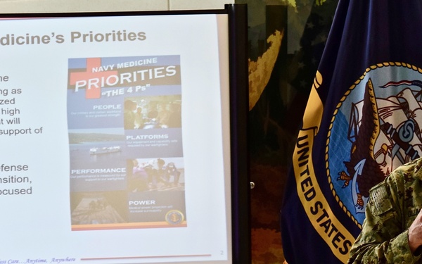 A U.S. Navy officer in camouflage uniform is presenting a briefing on 'Navy Medicine's Priorities' using a projected slideshow. The presentation outlines 'The 4 Ps' framework — People, Platforms, Performance, and Power — in support of maritime superiority. A U.S. Navy flag is visible in the background.