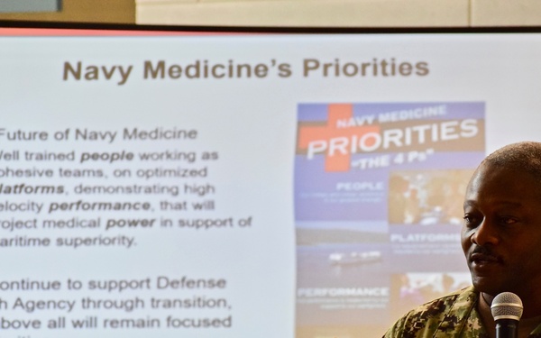 A U.S. Navy officer in camouflage uniform is presenting a briefing on 'Navy Medicine's Priorities' in front of a projected slide. The speaker, whose name tag reads 'SMITH,' is holding a microphone and gesturing while addressing an audience. The presentation outlines the future of Navy Medicine, emphasizing people, platforms, performance, and power.