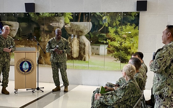 A formal military gathering is taking place in what appears to be the lobby of Naval Hospital Guam, identifiable by the podium seal and U.S. Navy flag. Several personnel in U.S. Navy NWU Type III (woodland camouflage) uniforms are present, with two individuals standing at a podium and microphone while others are seated or standing nearby. The event appears to be a briefing, ceremony, or official address.