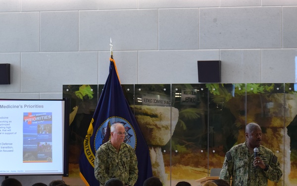 A military briefing or all-hands meeting is taking place in what appears to be a U.S. Navy facility. Two uniformed Navy personnel are presenting at the front of the room, with one speaking into a microphone while a slideshow titled 'Navy Medicine's Priorities' is displayed on a projection screen. A large audience of Navy personnel in camouflage uniforms are seated and attentive.