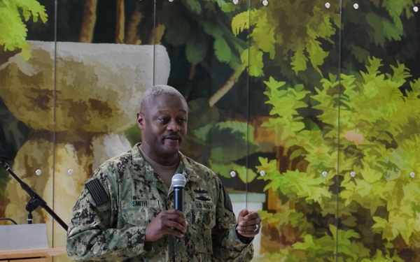 A U.S. Navy servicemember wearing NWU Type III (digital camouflage) uniform with the name tape 'SMITH' is addressing an audience of fellow military personnel while holding a microphone. The setting appears to be an indoor briefing or all-hands call at a Naval Hospital, as indicated by a partially visible seal on a podium reading 'NAVAL HOSPITAL G[UAM]'. Several uniformed personnel are seated in the foreground with their backs to the camera.