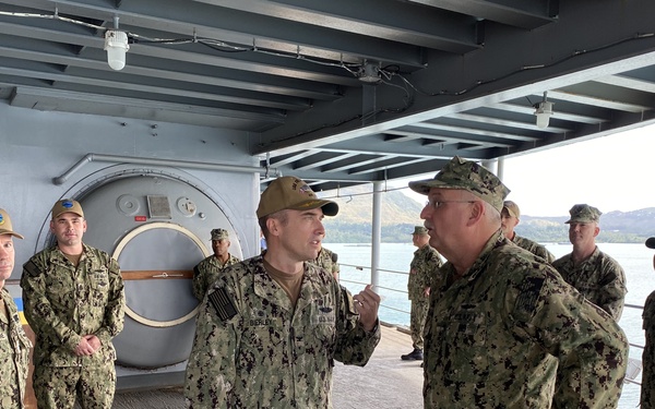A group of U.S. Navy personnel in NWU Type III (digital camouflage) uniforms are gathered on what appears to be the deck or hangar bay of a naval vessel. Two individuals in the foreground appear to be engaged in a briefing or discussion, with one sailor (name tape visible: 'BERLEY') addressing a senior officer. The setting appears to be a harbor or port environment, visible through the open side of the vessel.