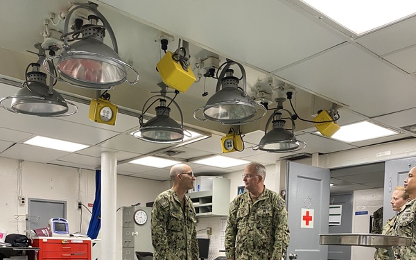 Military personnel in U.S. Navy camouflage uniforms (NWU Type III) are gathered inside what appears to be a shipboard medical/surgical facility. The room contains operating or examination tables, surgical lighting, and medical equipment consistent with a naval vessel's sick bay or operating room. A red cross symbol is visible on a gray door, confirming the medical nature of the space.