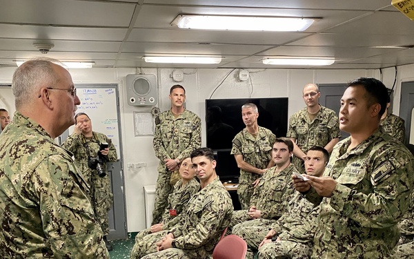 A group of U.S. Navy personnel in NWU Type III camouflage uniforms are gathered in what appears to be a briefing room aboard a naval vessel. A senior officer on the left is facing the group while a sailor on the right appears to be speaking or presenting. The setting includes institutional furnishings typical of a shipboard compartment.