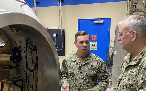 Two U.S. Navy personnel in NWU Type III camouflage uniforms are engaged in a discussion inside what appears to be a hyperbaric or pressure chamber facility. The younger sailor, identified by a name tape reading 'WRIGHT,' appears to be briefing or explaining something to the senior officer. A large cylindrical pressure chamber with gauges, valves, and cables is prominently visible to their left.