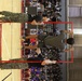 A gymnasium event appears to feature U.S. Marines in camouflage uniforms supervising or demonstrating a physical fitness activity using a red pull-up bar frame on a basketball court. Civilian participants, likely students, are attempting pull-ups or similar exercises on the red frame structure while a large audience of students watches from bleachers in the background. The event appears to be a school recruitment or fitness demonstration event.