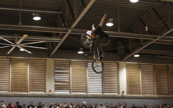 A BMX rider is performing a high aerial trick off a half-pipe ramp inside a gymnasium, with a large crowd of spectators watching from the bleachers. The event appears to be a Marines-sponsored BMX demonstration, as indicated by the 'Marines' branding and USMC Eagle, Globe, and Anchor insignia on the ramp. The setting is an indoor school or community gymnasium with industrial ceiling fans and overhead lighting.