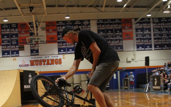 A BMX rider is performing a stunt trick on a gymnasium floor, balancing on the rear wheel of a black BMX bike. The event appears to be a school assembly or pep rally at a school called 'Mustangs,' with a large audience watching from the bleachers. A ramp is visible in the background, suggesting a planned stunt demonstration.