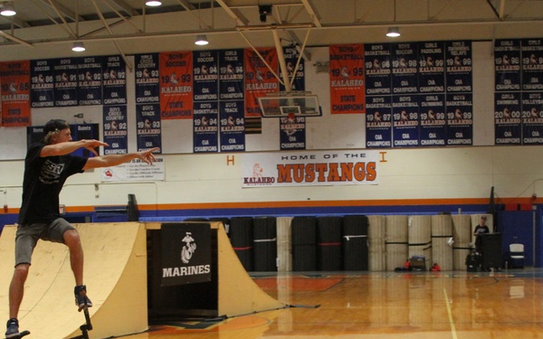 A BMX rider is performing a standing trick on his bike in a school gymnasium, balancing upright on the frame while extending his arms for balance. The event appears to be a sponsored demonstration by the U.S. Marines at Kalahe'o High School, home of the Mustangs. Championship banners from various years line the upper walls of the gymnasium.