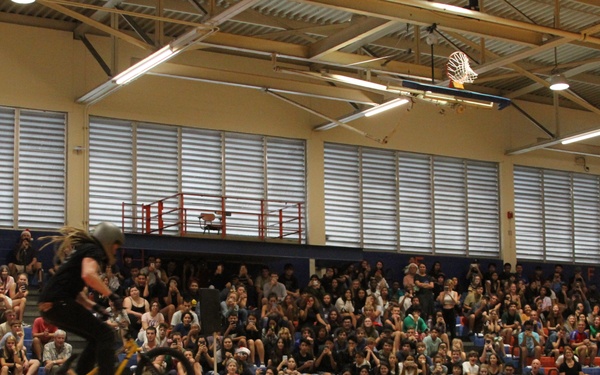 A BMX stunt rider is performing a jump over a row of people lying flat on a gymnasium floor during what appears to be a school assembly. The event is organized by ASA Action Sports High School Tour, with anti-bullying messaging visible on a banner. A large crowd of students fills the bleachers watching the performance.
