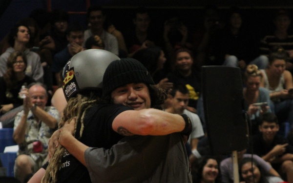 Two individuals are sharing an embrace at what appears to be a sporting event or competition held in an indoor arena. One person is wearing a helmet and the other a black beanie hat, suggesting a post-performance or post-competition moment. A large crowd of spectators is visible in the background, many appearing engaged and smiling.