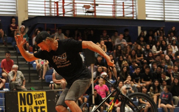 A BMX rider performs a trick on a single wheel (wheelie/manual) on a gymnasium basketball court during what appears to be a school assembly. The performer is balancing on the rear wheel only while a large crowd of students watches from bleachers on both sides. A 'No Place for Hate' Anti-Defamation League banner is prominently displayed in the background.