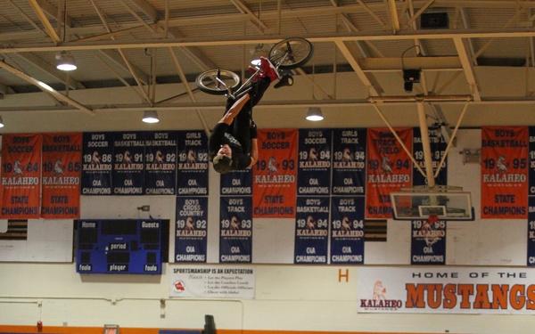 A BMX rider is performing an aerial trick inside the Kalaheo High School gymnasium, flipping upside down high above a wooden half-pipe ramp. The event appears to be a school assembly or demonstration, with spectators visible in the bleachers. A Marines-branded banner is displayed on the ramp, suggesting possible military recruitment sponsorship.