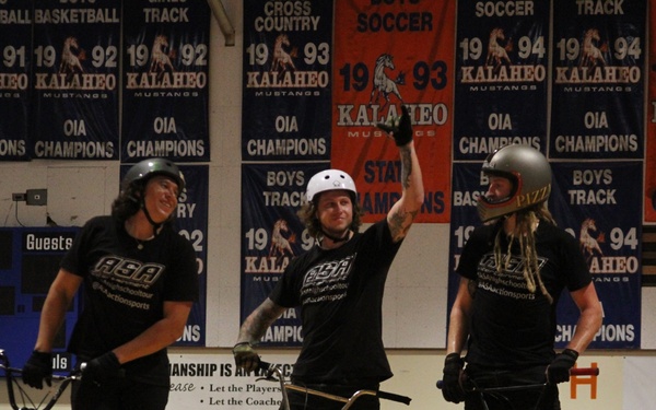 Three BMX riders wearing matching black 'ASA' (Action Sports Association) t-shirts and helmets are posed on their bikes atop a wooden ramp inside the Kalaheo Mustangs gymnasium. The center rider has one arm raised in a celebratory gesture. Championship banners from the 1980s-1990s for Kalaheo High School line the walls in the background.