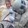 A military medic in ACU (Army Combat Uniform) and helmet is providing tactical combat casualty care (TCCC) to a simulated casualty with moulage (simulated wounds/blood) applied to the face. The medic is wearing blue nitrile gloves and appears to be assessing or treating an abdominal area, with a medical device visible on the casualty's torso. The scenario appears to be a training exercise based on the theatrical makeup used to simulate injuries.