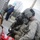 Two military personnel wearing advanced combat helmets with night vision mounts and ACU/multicam uniforms are attending to a simulated casualty near a chain-link fence. The scene appears to be a training exercise, as indicated by what appears to be a moulage (simulated wound makeup) on the casualty. Bystanders in civilian clothing are visible in the background near a green street sign.