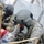 Two U.S. military personnel wearing flight helmets and camouflage uniforms are providing medical assistance to what appears to be a casualty during a training exercise or real operation. One soldier wears MultiCam/OCP pattern while the other wears the older ACU digital pattern, both equipped with aviation helmets. The scene includes simulated or actual blood visible on the casualty and surrounding materials, suggesting a medical response scenario.