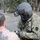 A military or National Guard personnel in full tactical gear, including a flight helmet with a GoPro camera mounted on it, is attending to a shirtless civilian male. The soldier is wearing MultiCam/OCP camouflage and has a Pennsylvania state patch visible on the right shoulder. The setting appears to be an outdoor area with bare trees and patchy snow, suggesting a cold-weather rescue or emergency response operation.